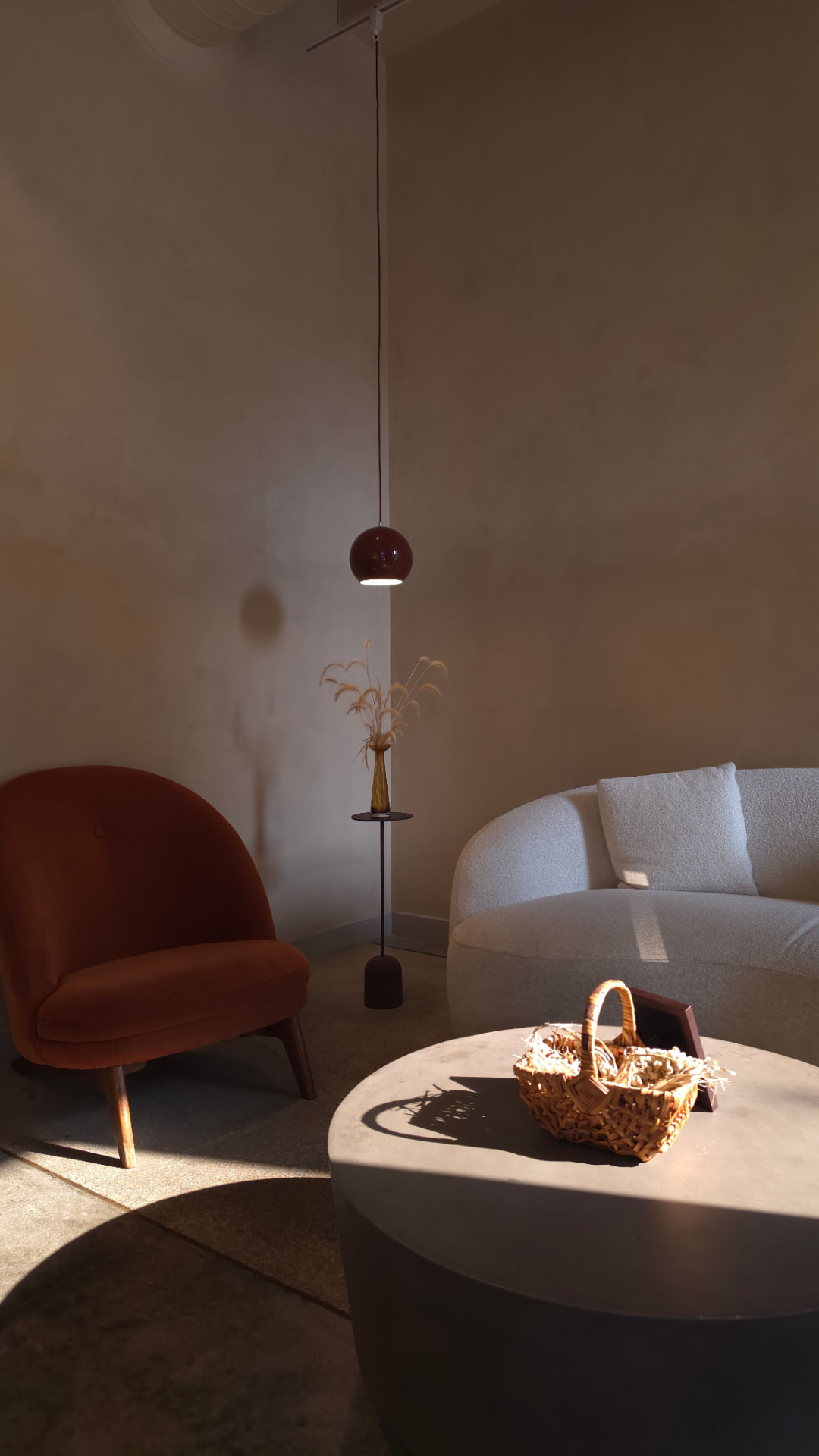 Zithoek met een terracottakleurige fauteuil, een crèmewitte gebogen bank, een bordeauxrode hanglamp en een ronde salontafel met een rieten mandje, in een warme beige ruimte met invallend zonlicht
